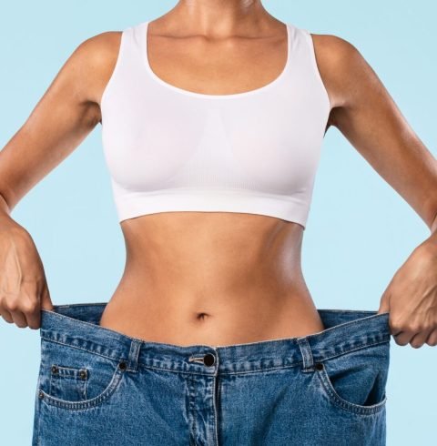 Closeup cropped view of skinny woman showing flat stomach pulling oversized big blue pants jeans. Slim body low fat. Healthy size athletic posing girl isolated over blue color studio background